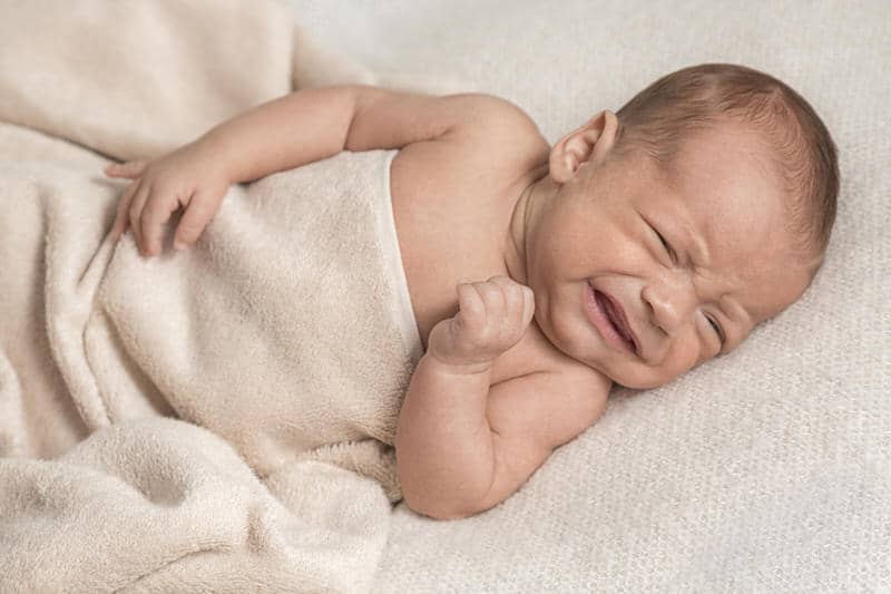 A newborn baby is laying down with a blanket and is crying.