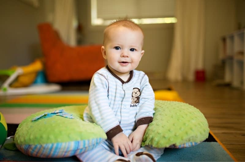 A young baby boy is able to sit up on his own with the support of a round pillow around his body.