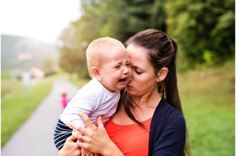 An infant boy is crying outside, while mom is holding him and trying to soothe him.