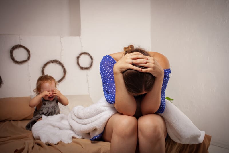 A tired mother is frustrated, while her toddler girl is sitting behind her crying. One of the negatives of extended breastfeeding is the continued reliance from the toddler on you to feed.