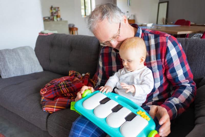 Grandpa is holding his infant grandson, sitting on the sofa. The little one isn't getting enough time to learn how to crawl on the floor, because he's always being held by someone.