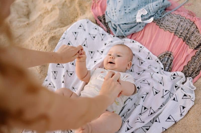 Mom has laid down her newborn baby on a blanket at the beach, to apply SPF on his skin.