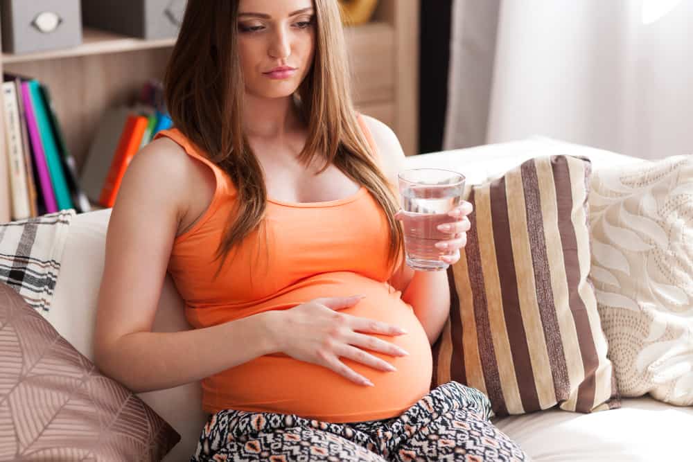 A young pregnant woman is sitting down and drinking water to help ease her nausea.