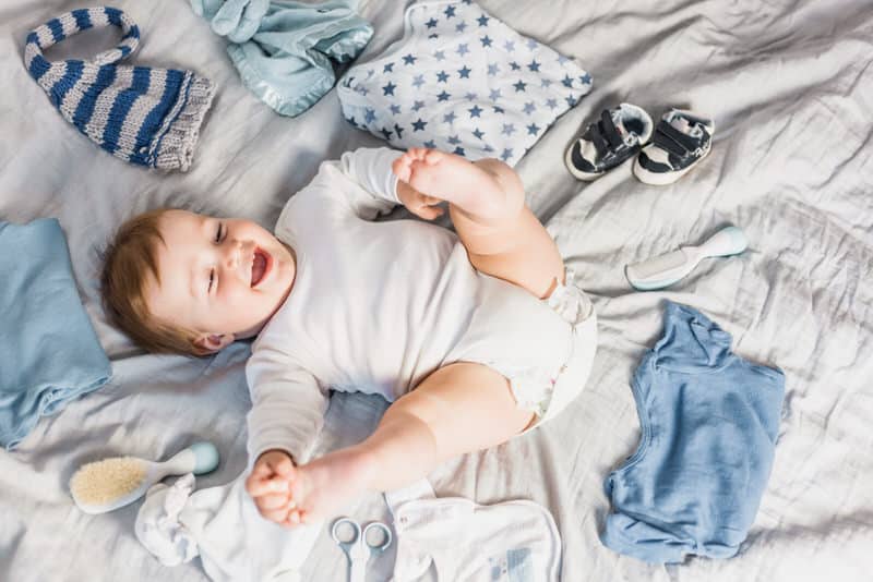 A happy infant boy is laying on his back in his crib, with different types of clothes surrounding him.