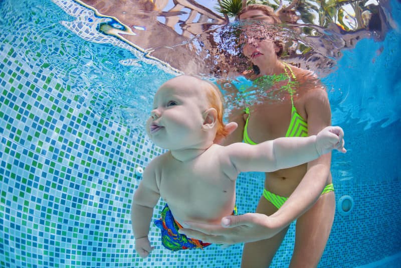 A swimming coach is teaching an infant boy how to swim underwater.
