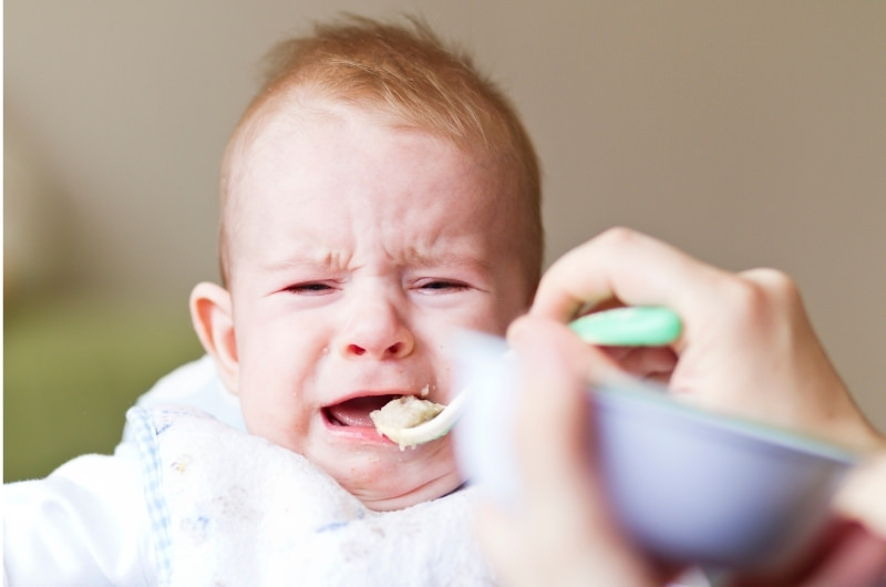 An infant boy is crying while sitting down and eating rice cereal.