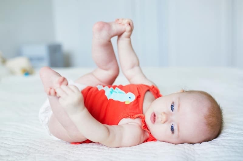 An infant girl is playing with her feet while laying down on her back.