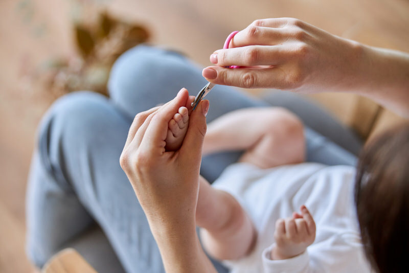 Mom has her newborn baby on her lap and is carefully cutting the newborn's toenails.