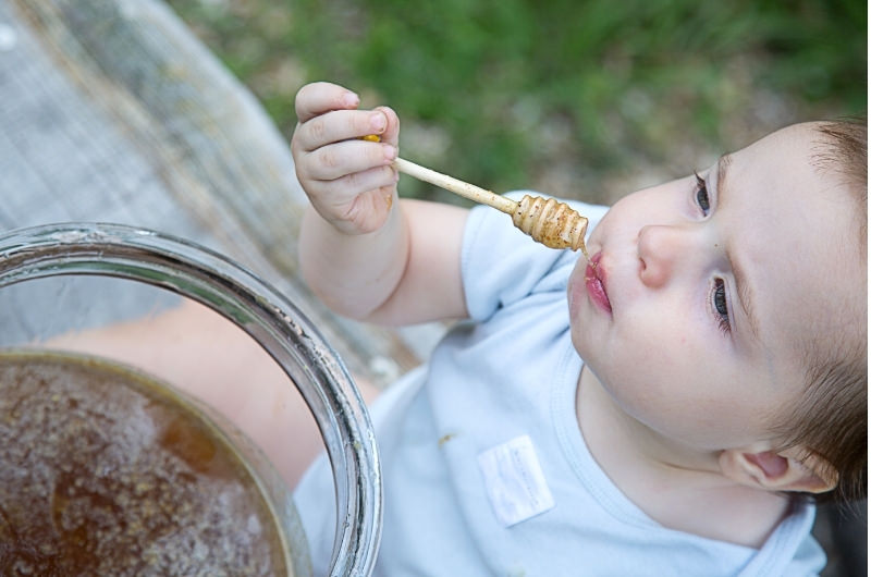 A toddler boy is sitting and tasting some honey for the first time, something he couldn't have when he was an infant or younger.