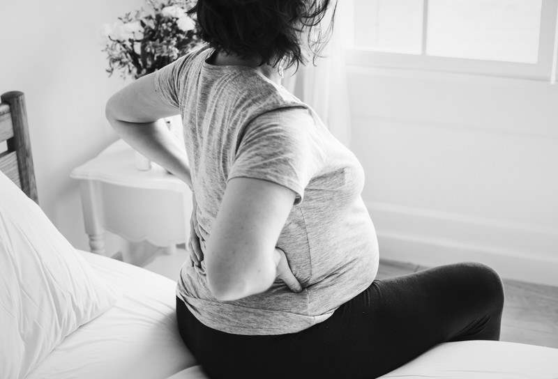 A young pregnant woman is sitting up on her bed in discomfort, possibly because she is eating too little with the one-a-day meal she's currently doing.