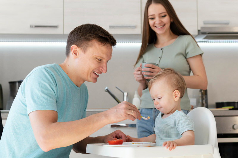 Dad and mom are helping feed their toddler son, who is sitting in a high chair.