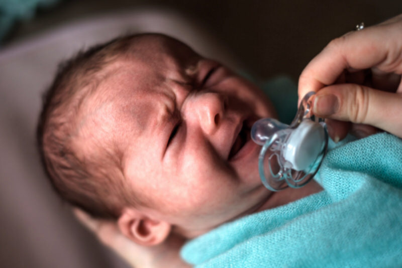 A newborn baby is crying and refusing to take a pacifier that's being offered by mom.