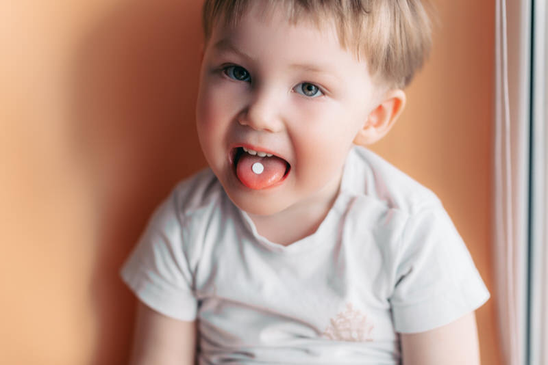 A toddler boy is sticking his tongue out and showing that he has a pill-shaped medicine in his mouth.