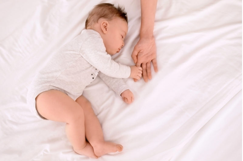 A 10-month infant boy is taking one of his daily naps on the bed while he holds his mommy's finger.