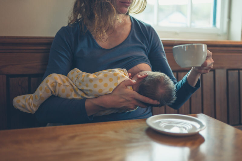 A mom is drinking coffee while breastfeeding her newborn son.