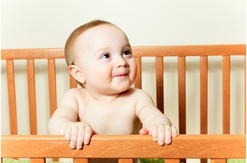 An infant boy is standing up inside his crib and happily looking upward at his parents.