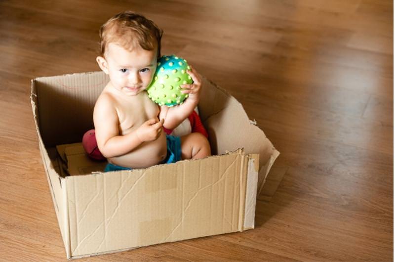 An infant boy is sitting in a cardboard box playing in there with some of his favorite toys.