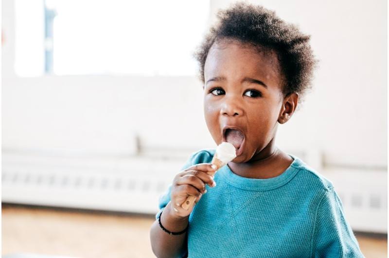 A toddler is happily snacking on some yogurt, too close to his next meal time, meaning he most likely won't eat much.