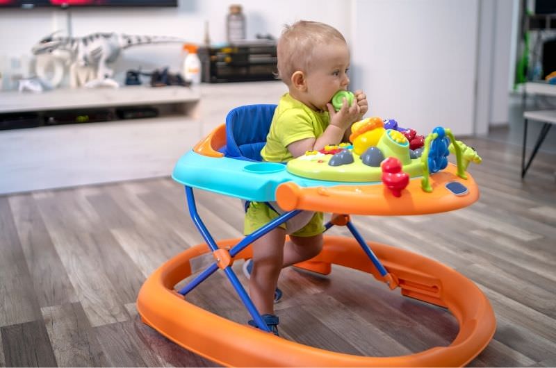 An infant boy is placed in a baby walker and is playing with toys and moving around the living room.