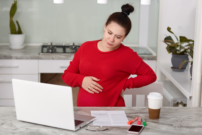 A pregnant woman is standing up in her kitchen, working on the island and having coffee.