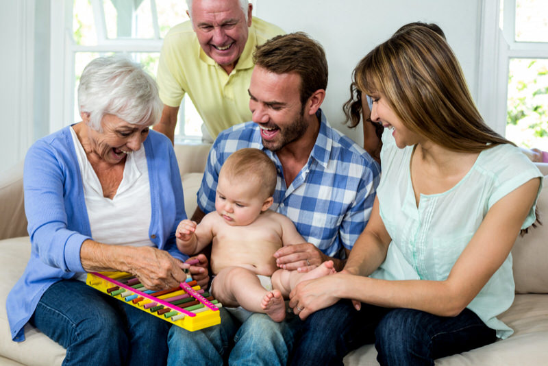 An infant boy is sitting with his parents and grandparents playing with a toy