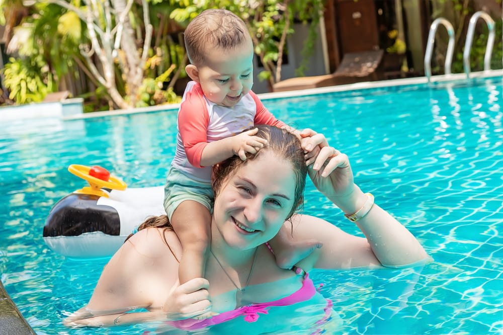 A toddler boy is in the pool sitting on his mom's shoulders and is pulling her hair