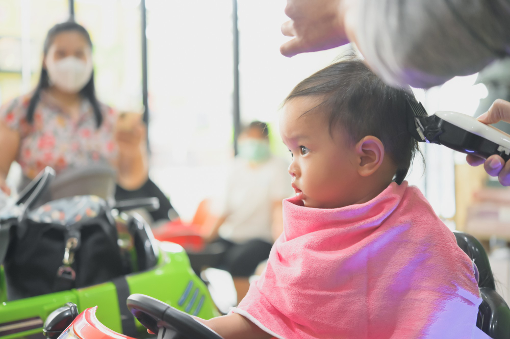 A baby girl is getting her first haircut at a salon while mom watches from the side