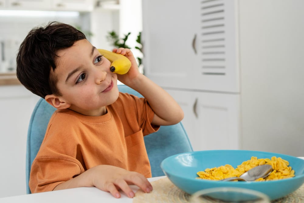 A toddler boy is sitting down eating his breakfast of cereal and a banana