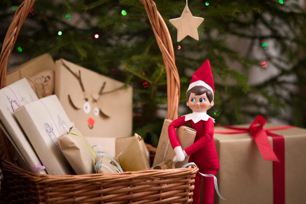 A toy elf is made to look like he's checking the gifts under the Christmas tree