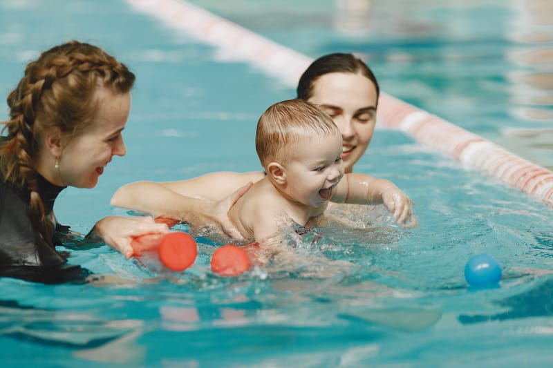 A toddler boy is learning how to swim with his mom and swimming instructor nearby