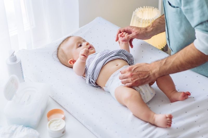 A dad lays down his toddler son on a changing table to start changing his diaper and treating his diaper rash with some ointment.