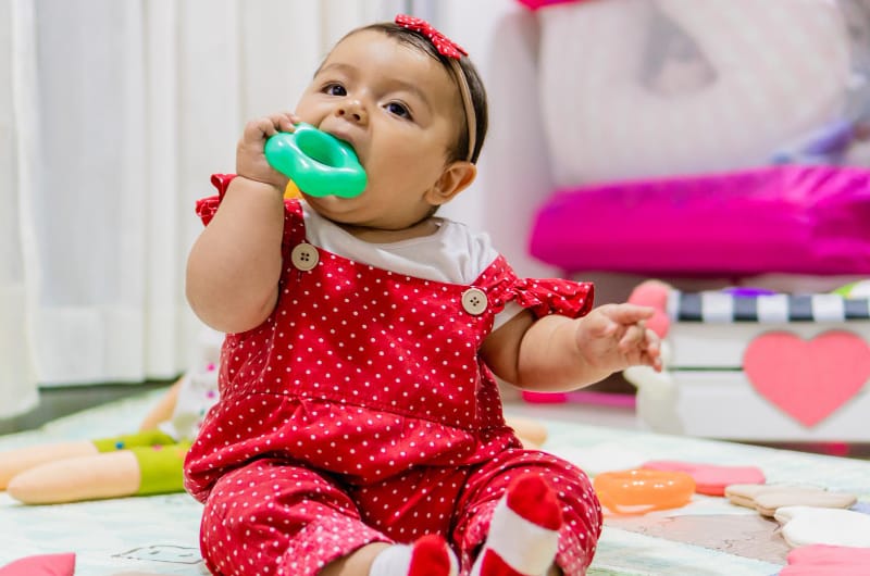 An infant girl is sitting up and chewing on a teething toy