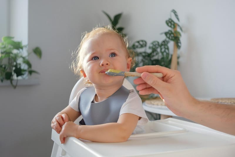 A toddler girl is sitting in her high chair, refusing to chew the food dad is spoon feeding her