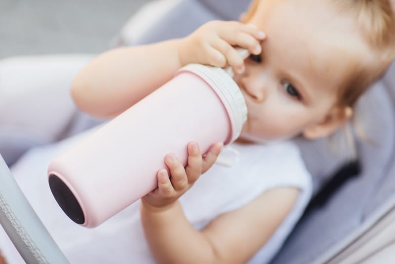 A toddler girl is holding a bottle on her own and drinking from it for the first time, hitting a new baby milestone.