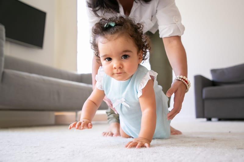 An infant girl is trying to crawl as mom watches her from behind