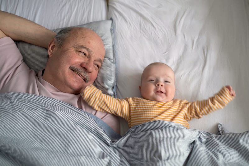 An infant boy is happily laying on the bed next to his grandfather