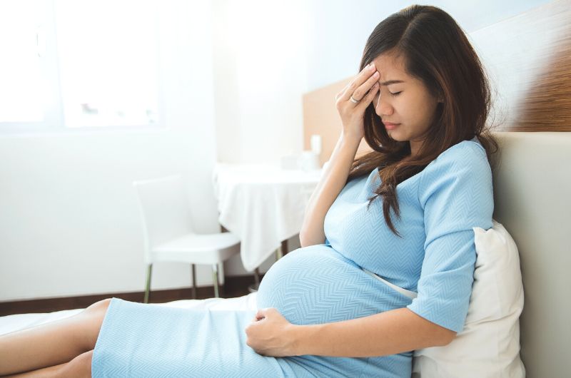A pregnant woman is sitting up on her bed and is noticeably not feeling well, she might have a fever.