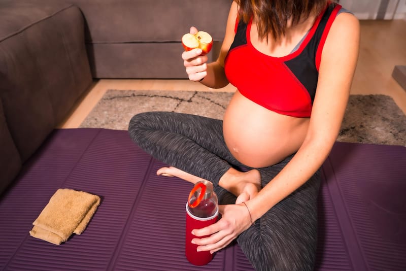 A pregnant woman is eating an apple after doing some light excersises on a yoga mat