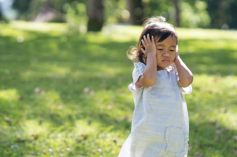 A toddler girl is covering her ears and not looking happy