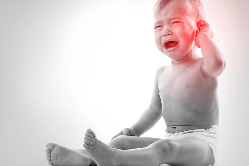 A toddler boy is sitting up and crying while touching his ear, because he might have an ear infection.