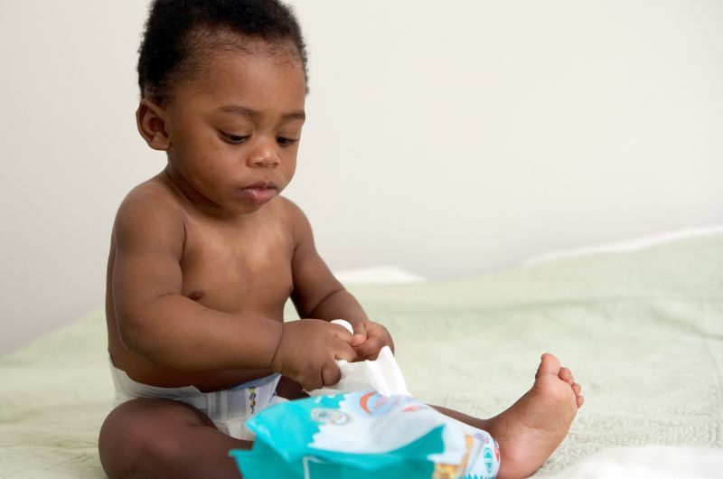 An infant boy is sitting up and is pulling a single wet wipe out of the container
