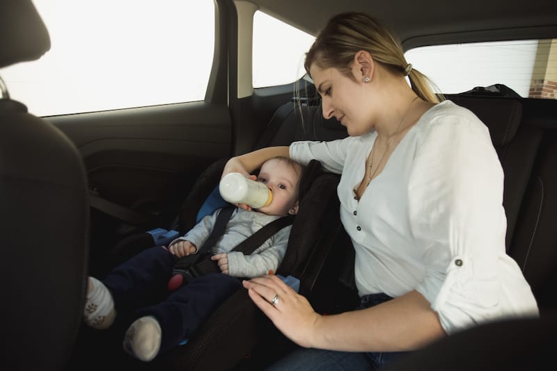 A mom is bottle-feeding her infant son in the back seat of a car