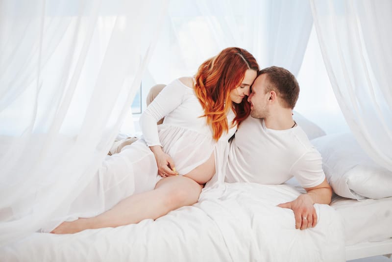 A young pregnant couple is in bed intimately hugging each other for a pregnancy photo