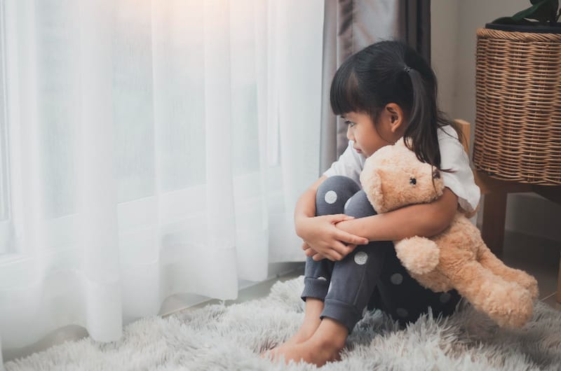 An anxious toddler girl is sitting down, hugging her teddy bear, and looking out the window to have some quiet time and calm down.