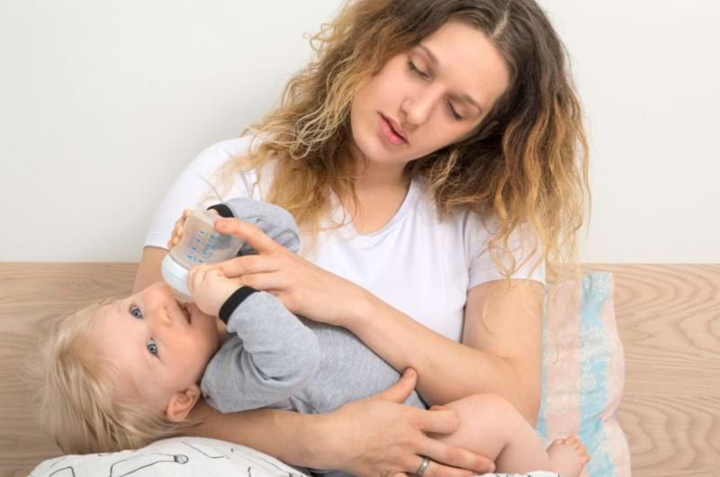 An exhausted mother is bottle-feeding her baby and forgetting to time the feeding.