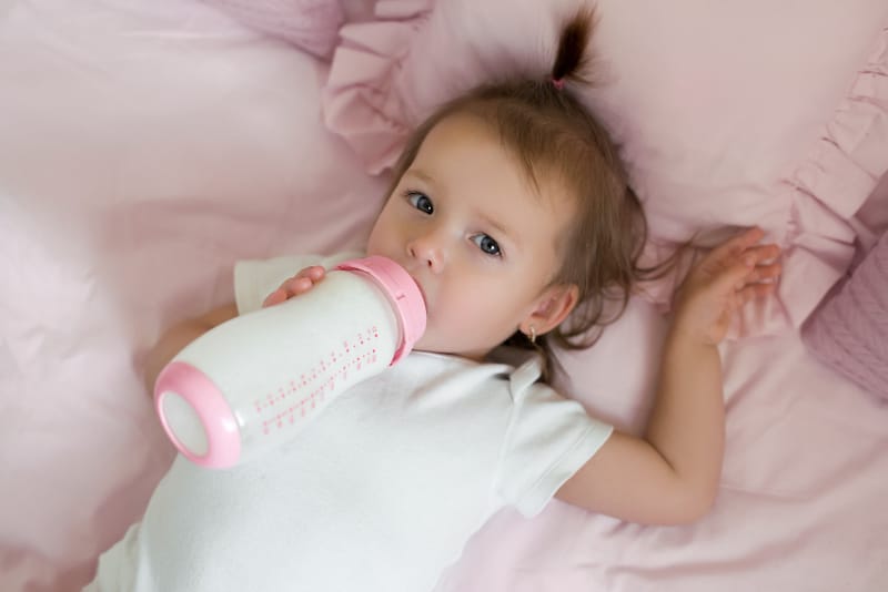 A toddler girl is lying down drinking whole milk from a baby bottle