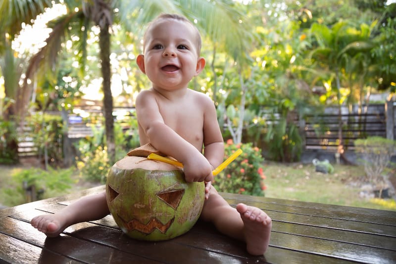 A baby boy is happily sitting down and drinking coconut water from a straw