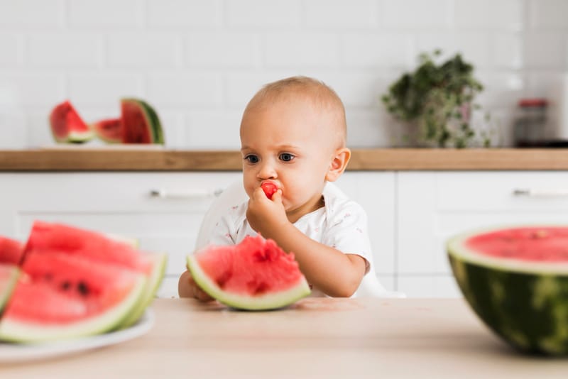 An infant boy is sitting up eating watermelon at the dining table