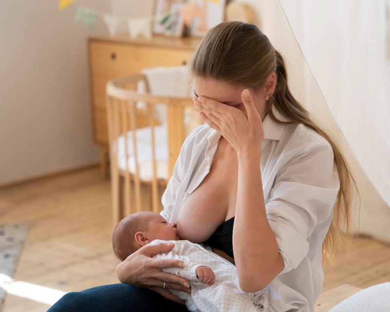 Mom is breastfeeding her baby, and she looks noticeably tense as she has one hand over her face