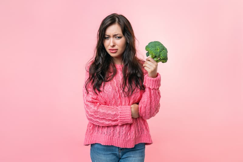 A breastfeeding mom is holding up a broccoli, thinking if it's good for her to eat it while she's breastfeeding, or if it might cause excess gas to her baby